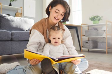 Genç anne, küçük çocuğuyla yerde oturup peri masalı okuyor. Bakıcı anne kitap tutuyor, bebeğiyle evde hikaye anlatmaktan zevk alıyor. Aile eğlence konsepti.