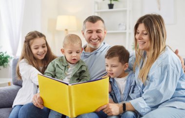 Beş çocuklu mutlu bir aile evde kanepede oturan çocuklarla kitap okuyor. Gülümseyen ebeveynler hafta sonunun tadını çıkaran çocuklarla vakit geçiriyorlar. İnsanlar ve aile eğlencesi konsepti.