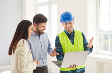 Mavi kasket ve güvenlik yeleği içinde gülümseyen inşaat işçisi parlak geniş evdeki genç aile çiftlerine dizüstü bilgisayardan bilgi gösteriyor. Mutlu adam ve kadın inşaatçıyla tasarım projesini tartışıyor..