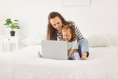 Anne ve küçük kızı dinleniyor, evdeki bir yatakta birlikte dinleniyorlar, internette dizüstü bilgisayar kullanarak online eğlence paylaşıyorlar. Aile bağlarını ve modern eğlenceleri ön plana çıkaran rahat bir an..
