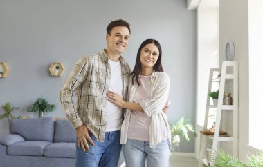 Evdeki oturma odasında dikilen ve sarılan genç çiftin portresi. İçerde kameraya bakan neşeli bir adam ve kadın. Aşk ve uyum ilişkileri kavramı.