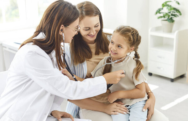 Little cute child girl with mother sitting in medical clinic with female family doctor. Pediatrician woman with stethoscope examining kids breathing and heartbeat at hospital. Health care concept.