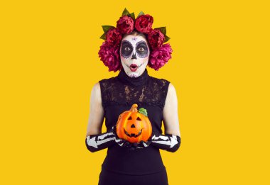 Cadılar Bayramı 'nda elinde balkabağı tutarken sana tuhaf bir şekilde bakan kadının portresi. Kafasında çiçek çelengi olan şeker kafalı kadın turuncu arka planda poz veriyor..