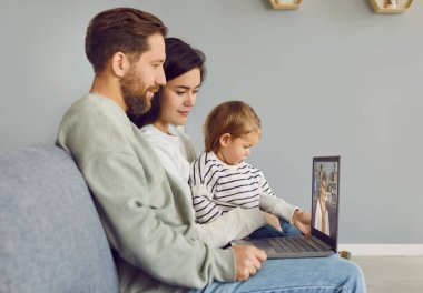 Evdeki oturma odasındaki kanepede oturan ve büyükannesiyle internetten video görüşmesi yapan mutlu, genç bir aile. Kızı olan çift dizüstü bilgisayar kullanıyor ve büyükannesiyle sohbet ediyor..