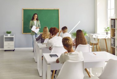 Bir grup çocuğun modern sınıflarda masalarda oturduğu bir ders, masa başında mutlu bir öğretmen, okuma yazma öğretiyor, çocuklara eğitim, gelişim bilgisi öğretiyor. 