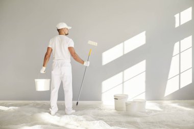 Ev yenileme servisinden bir ressam. Yeni ev ya da apartmanda çalışıyor. Arkadan bakan adam beyaz üniformalı, kovalı ve silindirli boya işçisi açık gri arka planda duruyor.