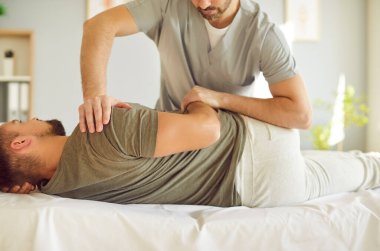 Profesyonel erkek kayropraktik uzmanı ya da osteopat uzmanı yetişkin bir adamla manuel terapi ofisinde çalışır. Osteopatik hekim, kontrollü hareketler kullanarak hastanın vücudunun üst kısmındaki ağrı kesicilere yardım ediyor..