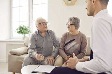 Gri saçlı yaşlı çift, finansal temsilciyle emeklilik sigortası hakkında konuşuyor. Evdeki koltukta oturuyor. Yaşlı adam ve kadın danışman ile bir araya gelerek yatırım sözleşmesi imzalayacaklar..