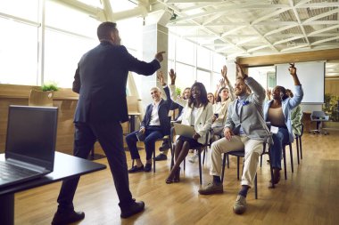Çeşitli aktif izleyiciler koçla iş eğitiminde sorular soruyorlar. Genç ve olgun çoklu etnik gruplardan oluşan bir grup ofiste oturmuş, gülümsüyor ve ellerini kaldırıyor.