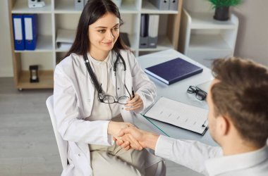 Klinikteki tıbbi muayeneden sonra masada oturan bir hastayla el sıkışan beyaz üniformalı, güler yüzlü, genç bir kadın doktor. İlaç ve sağlık hizmetleri kavramı.