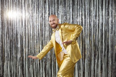 Neşeli adam altın bir kostüm içinde dans eder, dans eder, müzikten zevk alır ve bir partide parlak bir arka planda eğlenir. Kutlama, eğlence, disko havası ve enerjik bir akşam atmosferi içerir..