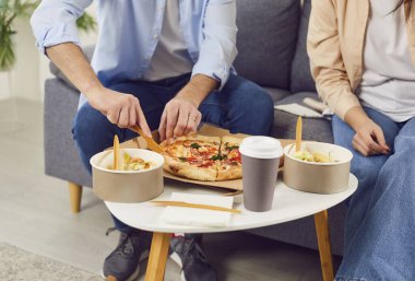Evdeki oturma odasındaki kanepede günlük kıyafetlerle oturan evli genç bir çift. Pizza, içki ve salata. Kadın ve erkek kanepede dinlenip fast food yiyorlar. Yemek siparişi ve teslimat konsepti.