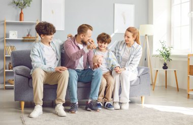 Çocuklu pozitif bir aile, evdeki domuz kumbarasına bozuk para koyup oturma odasındaki kanepede oturuyor. Tasarruf, mali planlama ve aile olarak zenginliği simgeliyor..