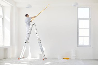 Boş bir odada merdivenin üzerinde duran ve tavanı boya silindiriyle boyayan üniformalı bir ressamın portresi. Yenileme ve yenileme konsepti.