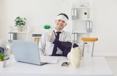 Bandajlı kolu ve bacağı kırık yakışıklı bir işadamı ofisinde oturuyor, çalışıyor ve başparmağını gösteriyor. Travma ve rehabilitasyon. Sosyal güvenlik ve sağlık sigortası kavramı.