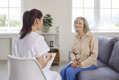 Genç, güler yüzlü bayan hemşire yaşlı, gri saçlı bir kadınla konuşuyor ve muayeneden sonra onu evinde ziyaret etmeyi öneriyor. Sağlık ve Bakımevi kavramı.