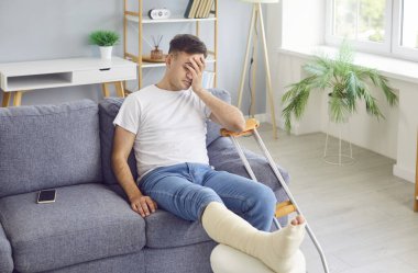 Kırık, bandajlı bacağı olan üzgün bir adam evdeki koltukta oturup acıdan kıvranıyor. Travma ve kırık da dahil olmak üzere yaraların tıbbi yönleri ve iyileşme sürecinde çekilen acıları vurgular..