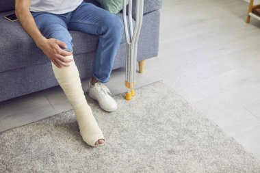 Kırık bir bacak ve koltuk değnekleriyle kanepede oturan bir adamın yakın çekim görüntüsü. Yaralanma ya da travma sonrası acı çekiyor. Yüzündeki ifade, iyileşmenin getirdiği rahatsızlığı ve zorlukları yansıtıyor..