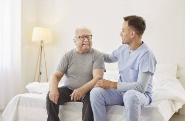 Dost canlısı genç erkek hemşire klinikte muayene ettikten sonra kıdemli bir adama danışıyor. Yaşlı, gri saçlı, hasta bir hasta hastanede yatağında bakıcı ile konuşuyor. Hemşirelik hizmeti kavramı.