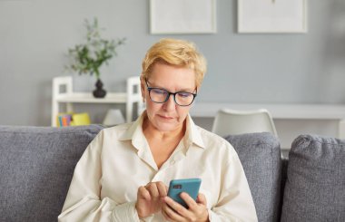 Yetişkin bir kadın akıllı telefon kullanıyor, uygulamalara bakıyor, evde oturuyor, telefon ekranında meşgul görünüyor, sohbet ediyor ya da internetten alışveriş yapıyor, boş zamanlarının tadını çıkarıyor yalnız başına dinleniyor, mesaj yazıyor, emekliliğin tadını çıkarıyor. 