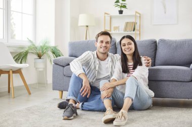 Oturma odasındaki yumuşak halıda birlikte oturan, kanepeye yaslanan ve huzurlu anın tadını çıkaran genç bir çiftin portresi. Mutlu adam ve kadın evde dinleniyor, sarılıyor ve kameraya bakıyor..