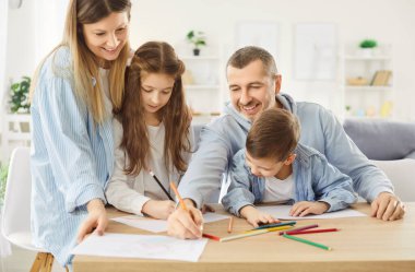 Küçük mutlu çocuklar evdeki oturma odasında neşeli aileleriyle masada oturuyor ve renkli kalemlerle resim çiziyorlar. Albümde resim yapan çocuklar aileleriyle vakit geçiriyor..