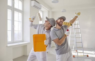 Profesyonel boyacılardan oluşan mutlu bir ekip ellerinde boya ruloları ile sanki mikrofonlar ve yüksek sesle şarkı söylüyor, tadilat yapıyor, beyaz duvarları ve merdivenleri olan boş bir odada poz veriyor.