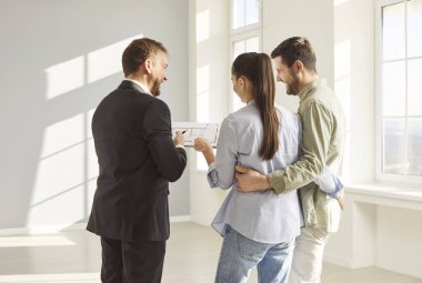 Takım elbiseli bir emlakçının parlak boş odada mutlu genç çifte daire düzeni sunuşunun arka görüntüsü. Gülümseyen adam ve kadın birbirlerine sarılıyorlar, gelecekteki ev alımlarını emlakçıyla tartışıyorlar..