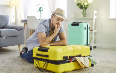 Mutlu gülümseyen adam evinde bavulunu toplayarak oturmuş internetten bilet ayırtan tablet ekran biletlerine bakıyor. Genç adam tatil gezisine hazırlanıyor. Tatil kavramı.