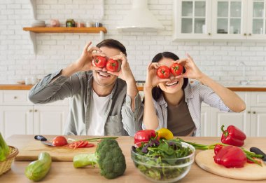 Mutlu ve eğlenceli bir çift modern mutfakta salata pişiriyor, gözlerinde taze domates sebzeleri tutuyor, gözlük yapıyor, ev aktivitelerinin keyfini çıkarıyor, sağlıklı organik gıda diyetini sürdürüyor.