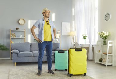 Mutlu gülümseyen adamın uzun bir portresi. Yaz tatiline hazır bavullarıyla oturma odasında duruyor. Neşeli erkek turist tatile çıkıyor. Seyahat ve seyahat kavramı.