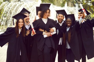 Gülümseyen mutlu uluslararası mezun öğrencilerin portresi üniversite mezuniyet elbisesi içinde eğleniyorlar ve açık havada diploma tutuyorlar. Eğitim ve mezuniyet kavramı.