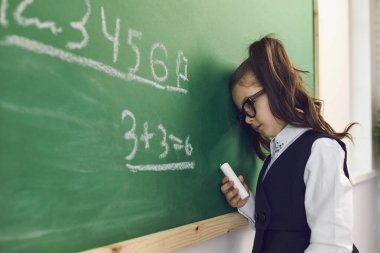 Gözlüklü, yeşil karatahtaya yaslanan ve matematik dersi sırasında hesap yapan bitkin bir kız.