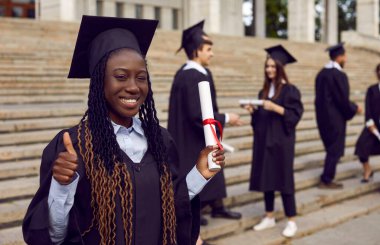 Genç, mutlu, gülümseyen, üniversite mezuniyet elbisesi giymiş, elinde diploması olan Afro-Amerikan bir kız öğrenci. Mezun kız arka planda sınıf arkadaşlarıyla dışarıda işaret veriyor..
