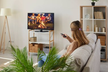 Televizyonda film izleyen bir kadın. Genç bayan rahat bir kanepede oturuyor ve televizyonunu değiştiriyor. Büyük kaliteli bir ekranı var. Modern rahat oturma odasının iç kısmında, duvarların üzerinde duruyor.