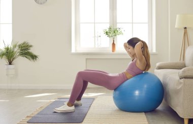 Evde aktif, narin genç bir kadın kaslarının kaslarını spor topu kullanarak çalıştırıyor. Spor antrenmanı sırasında ellerini başının arkasında tutan kızın spor yaparken sırt üstü yatışının yan görüntüsü..