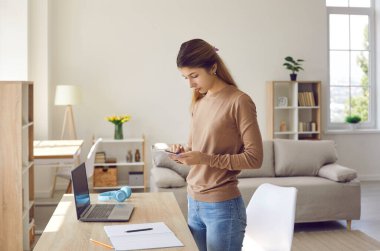 Cep telefonu kullanan bir kadın. Ciddi üniversite ya da üniversite öğrencisi, cep telefonu mesajlaşma uygulaması ya da uzaktan öğrenme platformu kullanarak, not alarak ya da web sözlüğüne danışarak, not defteri ile çalışma masasında duruyor