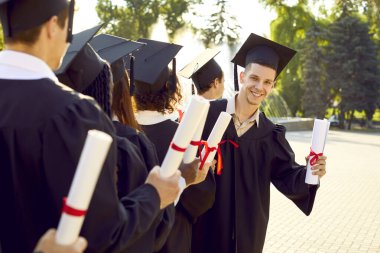 Mezuniyetinde mutlu üniversite, üniversite ya da liseli çocuk. Elinde diploması olan siyah şapkalı ve cüppeli genç adam diğer öğrencilerle sırada beklerken kameraya bakıp gülümsüyor.
