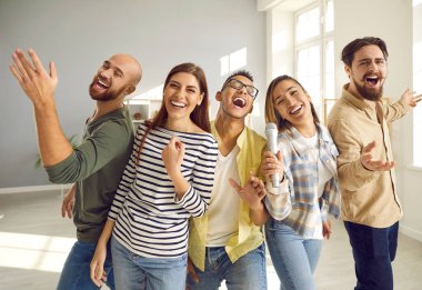 Genç, neşeli ve komik arkadaşları mikrofonla yüksek sesle şarkı söylüyor ve ev partisinde karaoke yaparken gülüyorlar. Gündelik giysiler içinde insanları yakın çekim. Arkadaşlık kavramı, ev eğlencesi ve insanlar.