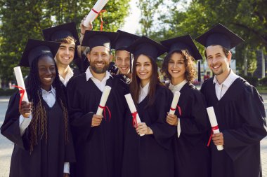 Mezuniyet gününde bir grup üniversite öğrencisi. Mezuniyet şapkalı ve cüppeli mutlu genç arkadaşlar yeşil kampüs bahçesinde duruyorlar, ellerinde diploma tomarları, kameraya bakıp gülümsüyorlar.