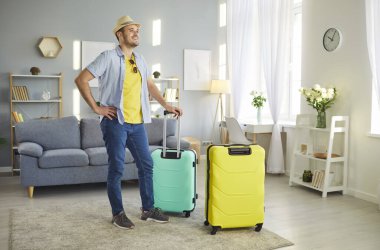 Sahil şapkalı mutlu genç adam yaz tatiline hazır bavullarıyla oturma odasında duruyor. Erkek turist tatile çıkıyor. Seyahat ve seyahat kavramı.