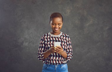 Mutlu koyu tenli kadın mesaj yazıyor ya da modern cep telefonunda sosyal ağlara takılıyor. Genç gülümseyen kadın gri arka planda duran yeni cihazının keyfini çıkarıyor. Modern cihazlar kavramı.