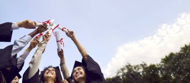 Bir grup gülümseyen mutlu mezun öğrencinin mezuniyet elbisesi içinde bir çemberin içinde diplomalarını gökyüzüne kaldırmalarının taban tablosu. Eğitim ve mezuniyet konsepti. Pankart.