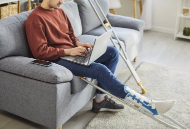 Kırık bacak ya da ayaktan yaralanan adam internette dizüstü bilgisayarla çalışıyor, koltuk değnekleriyle sargılı bacağı evde, rehabilitasyon sürecinde olan hasta, şişmiş ayak, üzerinde yürüyemeyen kişi, ciddi bir kırık.