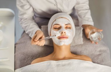 Güzellik salonundan güzellik kozmetolojisi prosedürü alan rahat, güzel bir kadının portresi. Kozmetik uzmanı klinikteki genç bir kız müşteriye nemlendirici maske takıyor. Cilt bakımı kavramı.