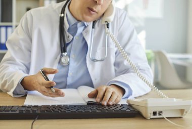 Ofisteki bilgisayarla çalışan, klinikteki hasta hastalara telefonla danışan, kendine güvenen, yüzü olmayan genç bir doktorun portresi. Tıp ve sağlık konsepti.