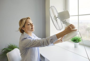 Elinde elektrikli vantilatör tutan genç ve gülümseyen bir kadın ofiste oturmuş serin havanın tadını çıkarıyor. Evde yaz sıcağından muzdarip neşeli bir kızın portresi. Soğutma ve sıcak sıcaklık kavramı.