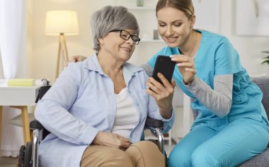 Tekerlekli sandalyedeki gülümseyen yaşlı kadın dost canlısı hemşireyle akıllı telefon kullanıyor. Neşeli kıdemli hasta, cep telefonunda bakıcıyla komik bir şey paylaşıyor. Bakımevi servisi ve hasta desteği.