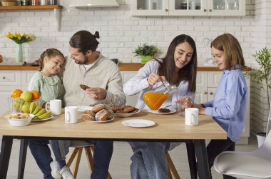 Birlikte kahvaltı yapmak, meyve suyu ile lezzetli hamur işleri yemek, mutfak içlerine karşı masada poz vermek için ünlü bir şekilde toplandık. Anne, baba, kız ve oğul evde lezzetli bir şeyler yiyorlar.