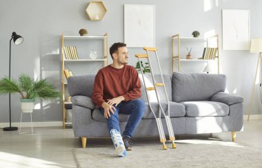 Hasta olduğu sırada evinde dinlenen bacağından sakatlanan genç bir adam. Bileği kırık üzgün bir adam koltukta koltuk değnekleriyle oturuyor.